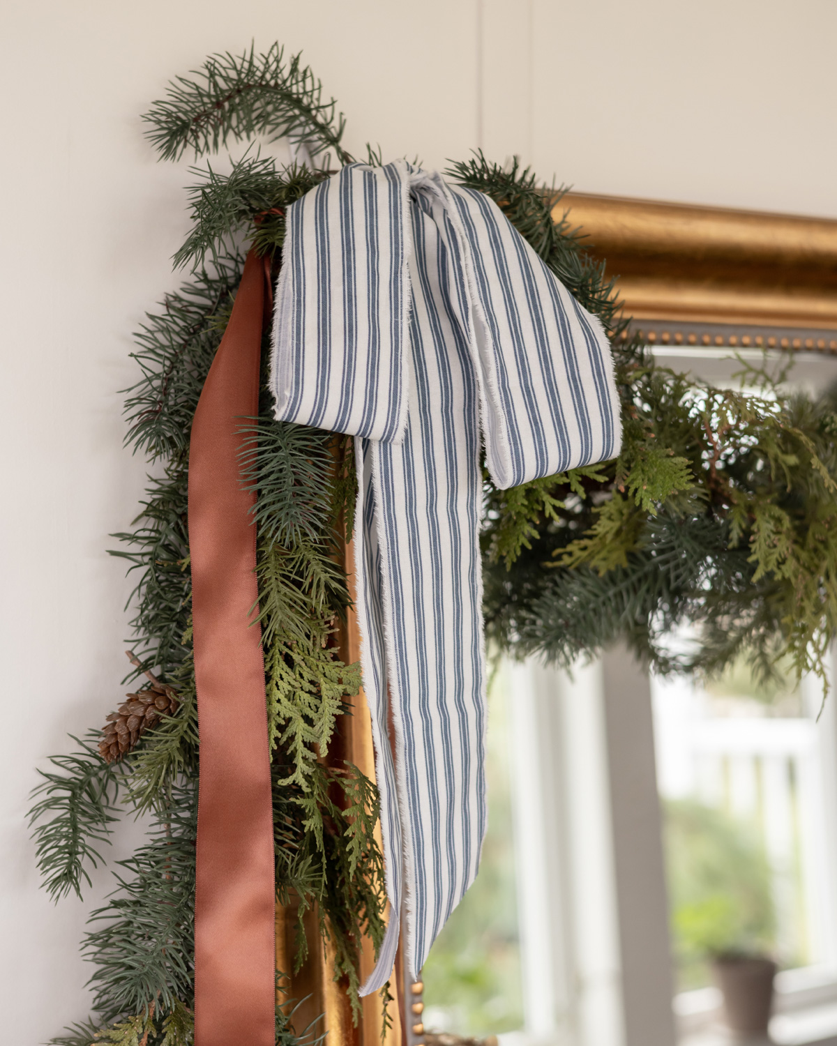 A garland hung over a mirror with faux garland, fresh greenery, and a blue striped bow.