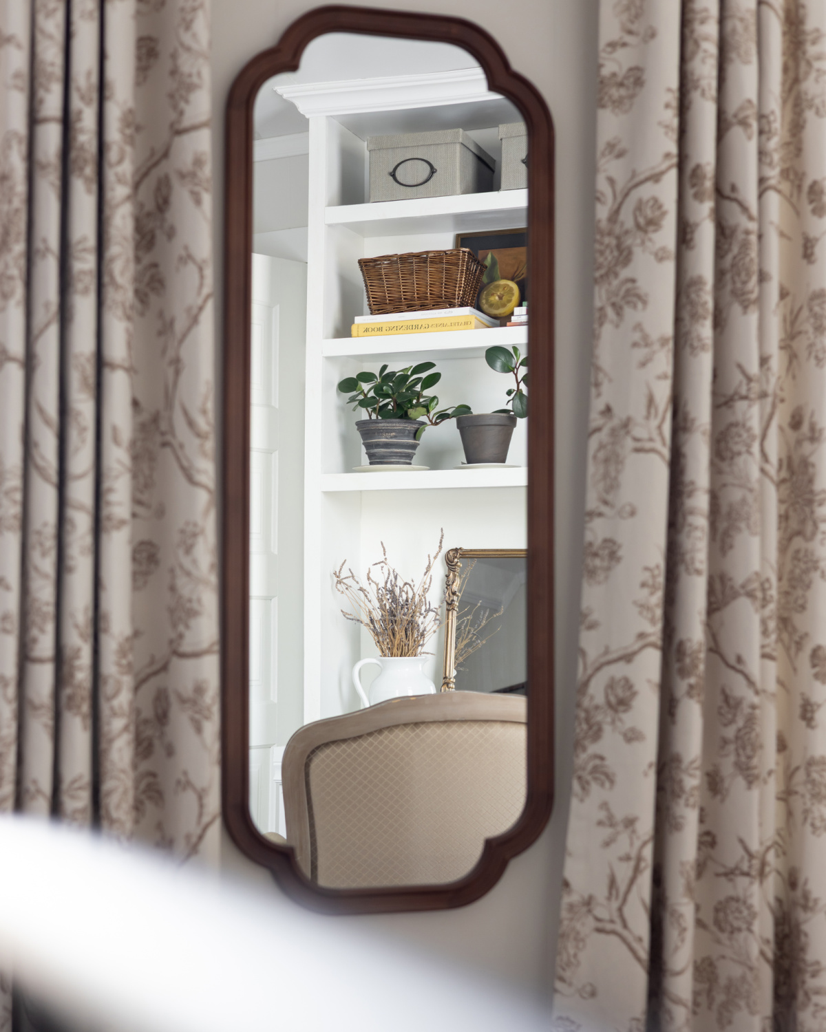 A built-in bookshelf is reflected in a mirror between floral curtains.